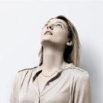 Frau mit hellbraunem Haar und beiger Bluse blickt nach oben und steht vor einem schlichten weißen Hintergrund.