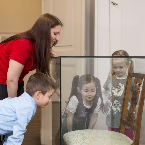 Eine Frau und drei kleine Kinder betrachten einen Holzstuhl, der in einem Museum oder einer Galerie hinter Glas ausgestellt ist.