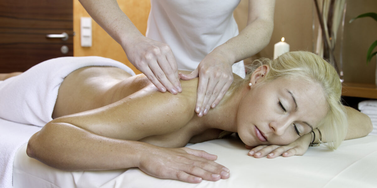 Eine Frau liegt mit dem Gesicht nach unten auf einem Massagetisch und hat die Augen geschlossen, während eine Masseurin ihre Schultern massiert.