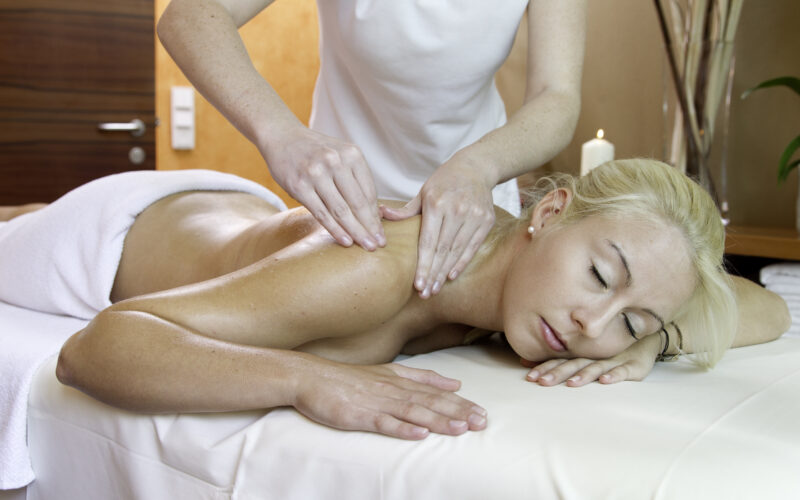 Eine Frau liegt mit dem Gesicht nach unten auf einem Massagetisch und hat die Augen geschlossen, während eine Masseurin ihre Schultern massiert.