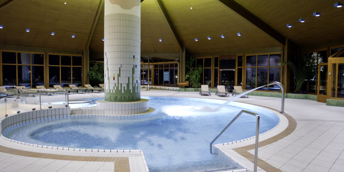 Innenpool mit zentraler gefliester Säule, Liegestühlen, Wasserstrahl und Holzdecke, umgeben von großen Fenstern und Topfpflanzen.