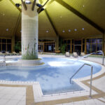 Innenpool mit zentraler gefliester Säule, Liegestühlen, Wasserstrahl und Holzdecke, umgeben von großen Fenstern und Topfpflanzen.