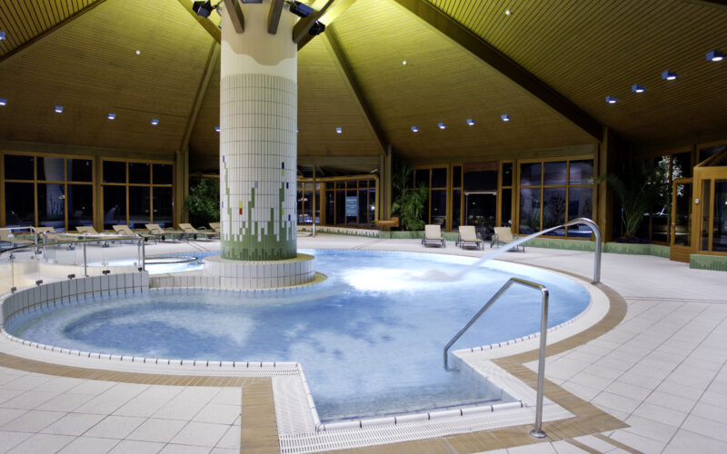 Innenpool mit zentraler gefliester Säule, Liegestühlen, Wasserstrahl und Holzdecke, umgeben von großen Fenstern und Topfpflanzen.