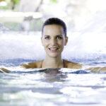 Eine Frau mit nassem Haar lächelt, während sie in einem Schwimmbad steht, die Arme ausgestreckt hat und das Wasser im Hintergrund plätschert.