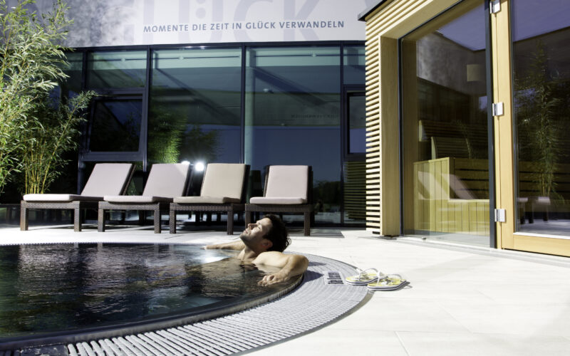 Ein Mann entspannt sich in einem runden Außenpool neben mehreren Liegestühlen und einem modernen Gebäude mit großen Glasfenstern.