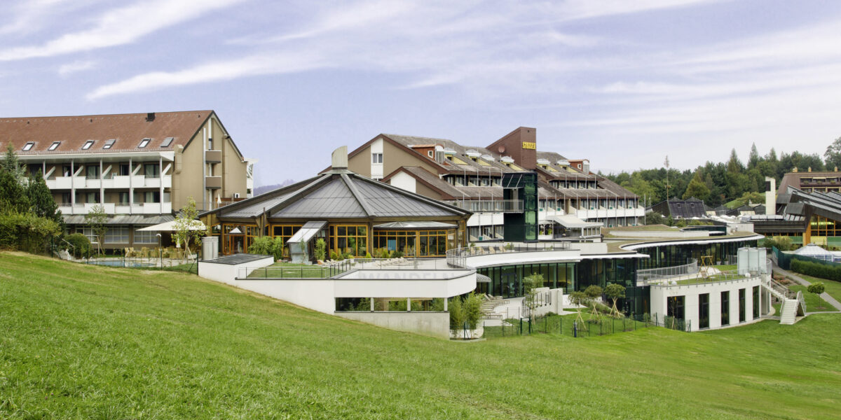 Ein moderner Hotelkomplex mit mehreren miteinander verbundenen Gebäuden und großen Fenstern, der auf einem grasbewachsenen Hügel unter einem teilweise bewölkten Himmel liegt.