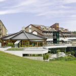 Ein moderner Hotelkomplex mit mehreren miteinander verbundenen Gebäuden und großen Fenstern, der auf einem grasbewachsenen Hügel unter einem teilweise bewölkten Himmel liegt.