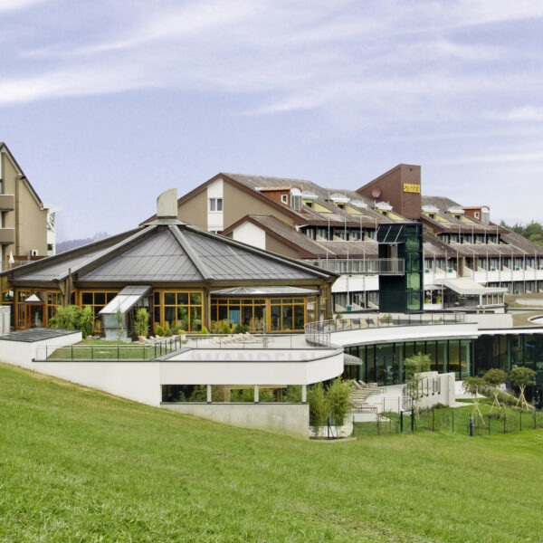 Ein moderner Hotelkomplex mit mehreren miteinander verbundenen Gebäuden und großen Fenstern, der auf einem grasbewachsenen Hügel unter einem teilweise bewölkten Himmel liegt.