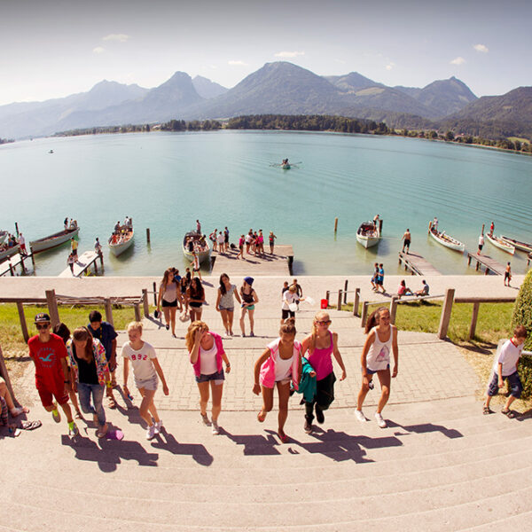 Eine Gruppe von Menschen geht eine Treppe von einem Steg am See hinauf, mit Booten auf dem Wasser und Bergen im Hintergrund unter einem klaren Himmel.