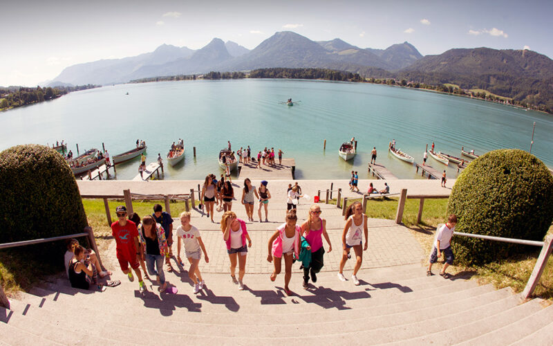 Eine Gruppe von Menschen geht eine Treppe von einem Steg am See hinauf, mit Booten auf dem Wasser und Bergen im Hintergrund unter einem klaren Himmel.