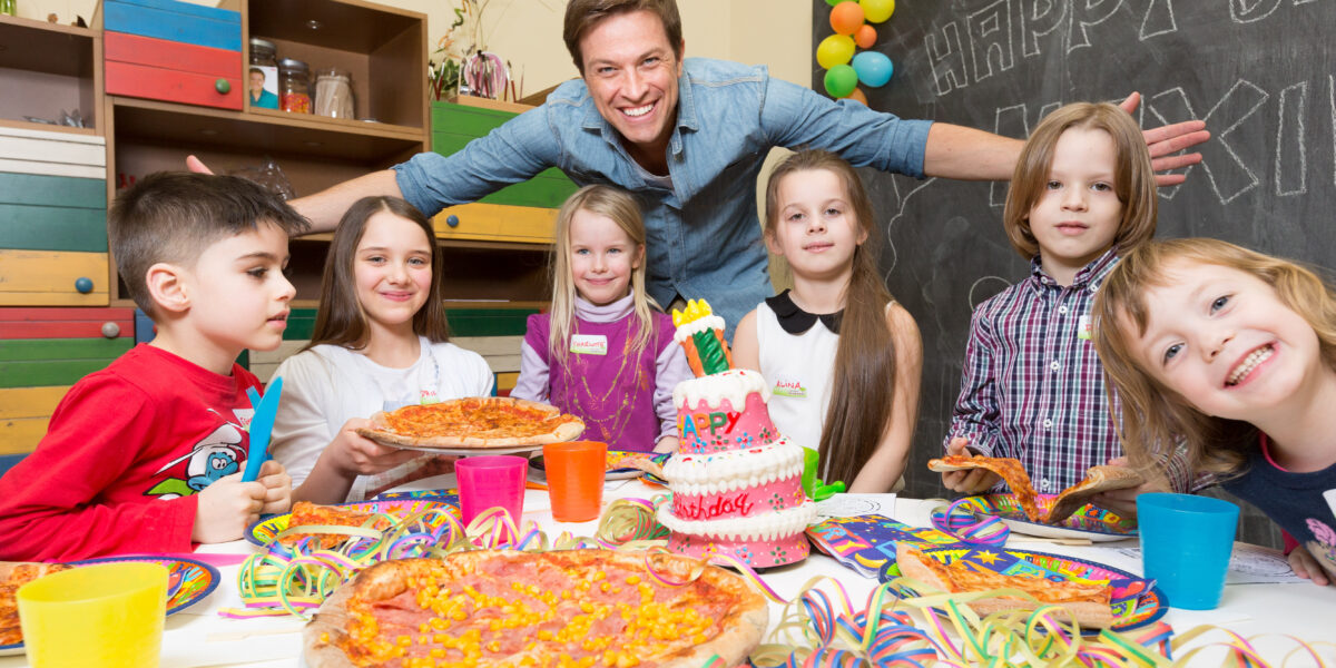 Eine Gruppe von Kindern und ein Erwachsener versammeln sich bei einer Geburtstagsparty um einen Tisch mit Pizza, einer Geburtstagstorte, bunten Bechern und Dekorationen.