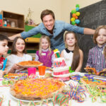 Eine Gruppe von Kindern und ein Erwachsener versammeln sich bei einer Geburtstagsparty um einen Tisch mit Pizza, einer Geburtstagstorte, bunten Bechern und Dekorationen.