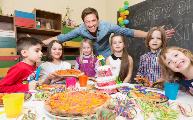 Eine Gruppe von Kindern und ein Erwachsener versammeln sich bei einer Geburtstagsparty um einen Tisch mit Pizza, einer Geburtstagstorte, bunten Bechern und Dekorationen.
