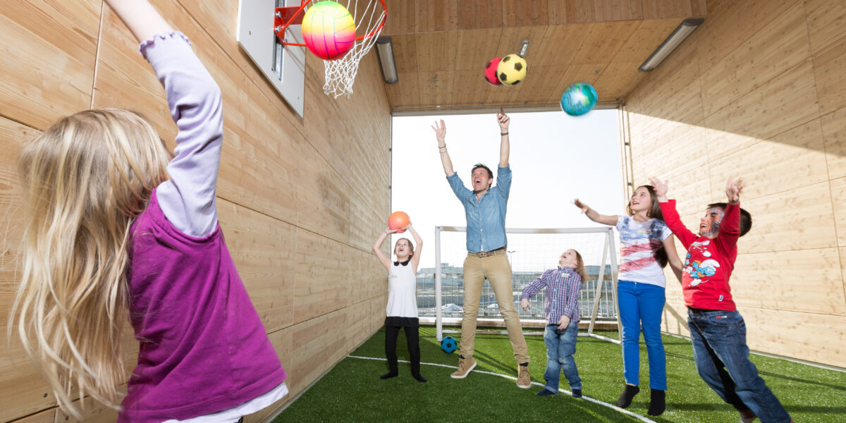 Eine Gruppe von Kindern und ein Erwachsener spielen Basketball und werfen Bälle in einer hölzernen Sporthalle mit Kunstrasen.