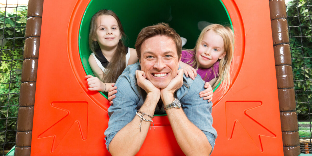 Ein erwachsener Mann und zwei junge Mädchen lächeln in die Kamera, während sie aus einer kreisförmigen Tunnelstruktur auf einem Spielplatz lehnen.