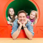 Ein erwachsener Mann und zwei junge Mädchen lächeln in die Kamera, während sie aus einer kreisförmigen Tunnelstruktur auf einem Spielplatz lehnen.