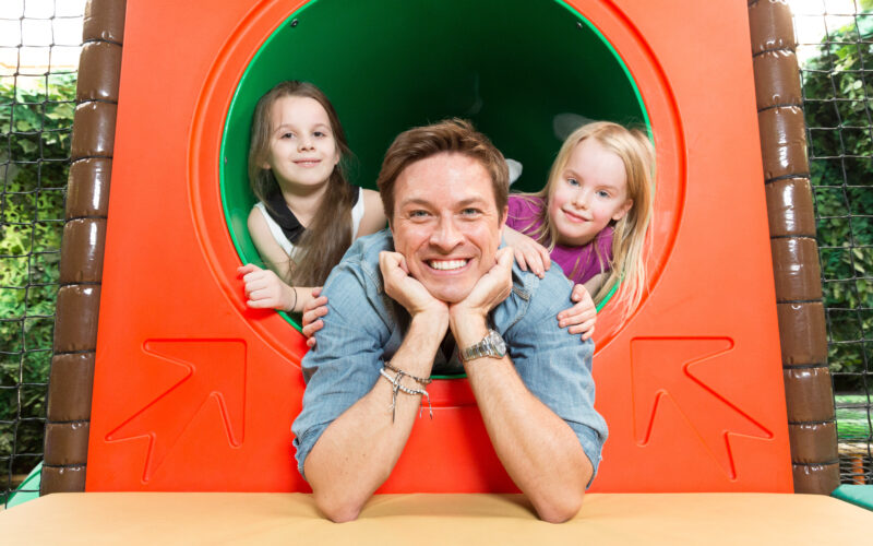 Ein erwachsener Mann und zwei junge Mädchen lächeln in die Kamera, während sie aus einer kreisförmigen Tunnelstruktur auf einem Spielplatz lehnen.