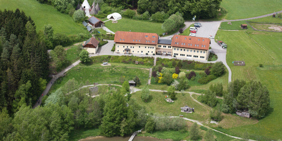 Luftaufnahme eines großen gelben Gebäudes mit rotem Dach, umgeben von Bäumen, offenen Feldern, geparkten Autos, Zelten und einem Steg, der in einen See hineinragt.