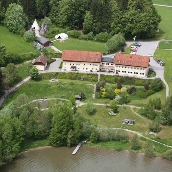 Luftaufnahme eines großen gelben Gebäudes mit rotem Dach, umgeben von Bäumen, offenen Feldern, geparkten Autos, Zelten und einem Steg, der in einen See hineinragt.