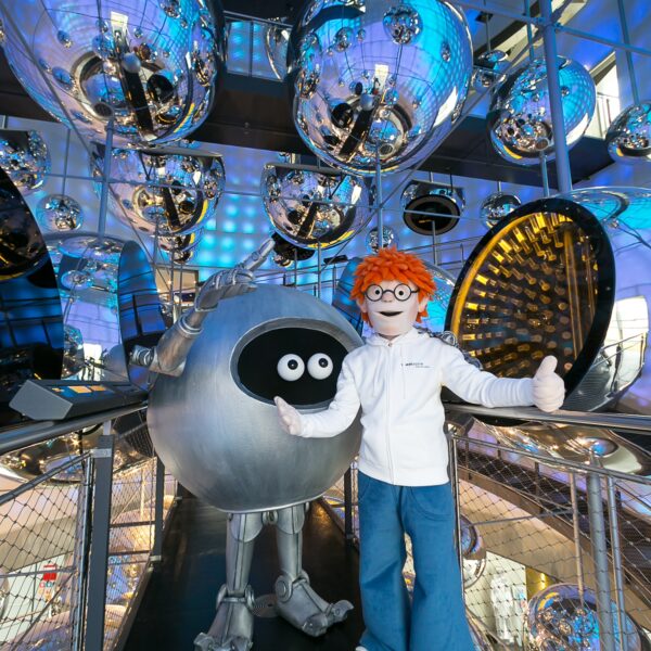 Eine Person in einem rothaarigen Maskottchen-Kostüm steht neben einer großen, runden Roboterfigur, umgeben von reflektierenden silbernen Kugeln in einer modernen, blau beleuchteten Innenumgebung.
