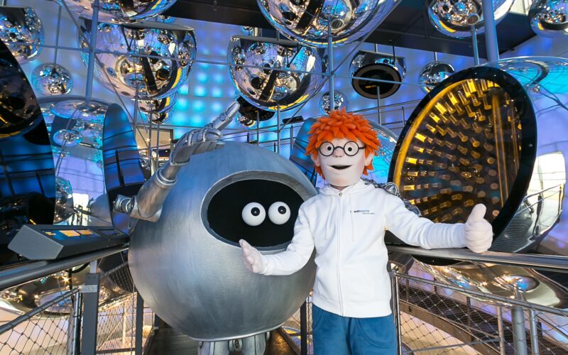 Eine Person in einem rothaarigen Maskottchen-Kostüm steht neben einer großen, runden Roboterfigur, umgeben von reflektierenden silbernen Kugeln in einer modernen, blau beleuchteten Innenumgebung.