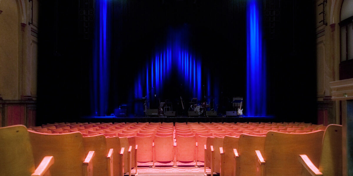 Leeres Theater mit Sitzreihen vor einer mit blauem Licht beleuchteten Bühne, auf der Musikinstrumente und -geräte zu sehen sind.