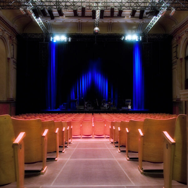 Leeres Theater mit Sitzreihen vor einer mit blauem Licht beleuchteten Bühne, auf der Musikinstrumente und -geräte zu sehen sind.