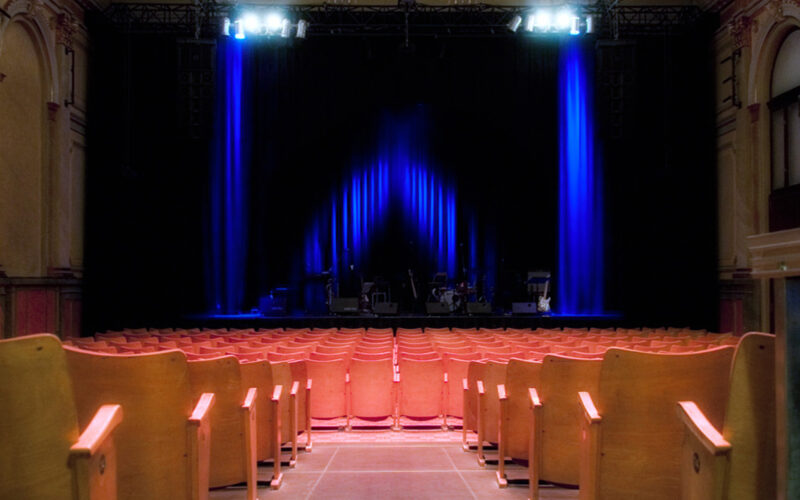 Leeres Theater mit Sitzreihen vor einer mit blauem Licht beleuchteten Bühne, auf der Musikinstrumente und -geräte zu sehen sind.