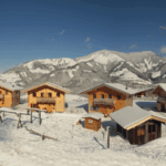 Die Holzhütten sind von Schnee umgeben, mit einer Bergkette im Hintergrund und einem strahlend blauen Himmel.