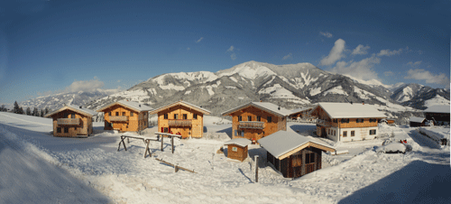 Die Holzhütten sind von Schnee umgeben, mit einer Bergkette im Hintergrund und einem strahlend blauen Himmel.