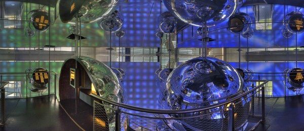 Ein futuristischer Raum mit metallischen Gängen, großen reflektierenden Kugeln, Netzgeländern und blau und grün beleuchteten Wänden, die eine verspiegelte, immersive Umgebung schaffen.