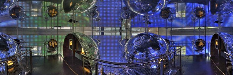 Ein futuristischer Raum mit metallischen Gängen, großen reflektierenden Kugeln, Netzgeländern und blau und grün beleuchteten Wänden, die eine verspiegelte, immersive Umgebung schaffen.