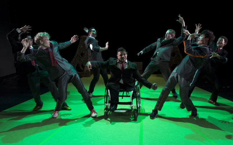 Eine Gruppe von Darstellern in Anzügen, darunter eine Person im Rollstuhl, posiert dramatisch auf einer grün beleuchteten Bühne.