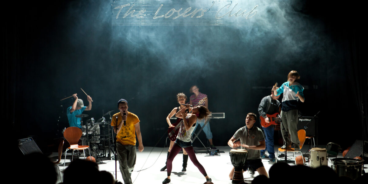 Eine Band spielt energisch auf der Bühne unter dramatischer Beleuchtung, mit verschiedenen Instrumenten und einem Schild mit der Aufschrift "The Lovers Club" im Hintergrund.