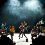 Eine Band spielt energisch auf der Bühne unter dramatischer Beleuchtung, mit verschiedenen Instrumenten und einem Schild mit der Aufschrift "The Lovers Club" im Hintergrund.