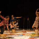 Vier Personen treten energisch auf der Bühne auf, balancieren auf kleinen Hockern, tragen bunte Outfits und einen gemusterten Boden vor einem dunklen Hintergrund.