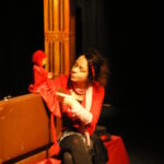 Eine rot gekleidete Frau sitzt neben einer braunen Schachtel und interagiert mit einer roten Marionette. Im Hintergrund ist ein hohes Gebäude mit einer Uhr zu sehen.