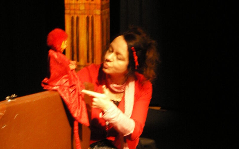 Eine rot gekleidete Frau sitzt neben einer braunen Schachtel und interagiert mit einer roten Marionette. Im Hintergrund ist ein hohes Gebäude mit einer Uhr zu sehen.