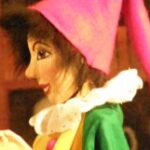 Nahaufnahme einer Marionette oder Puppe mit bemaltem Gesicht, die einen hellrosa spitzen Hut, ein grünes Kleid und einen weißen Rüschenkragen trägt.