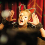 Person in einem Katzenkostüm mit Gesichtsbemalung und roten Ohren, die vor roten Vorhängen einen verspielten Ausdruck macht.