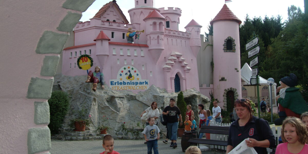 Menschen, darunter Kinder und Erwachsene, laufen und spielen in der Nähe eines rosafarbenen Märchenschlosses im Erlebnispark Strasswalchen. Im Hintergrund sind Schilder und Grünflächen zu sehen.