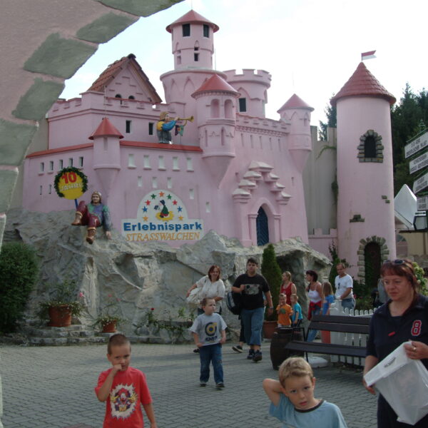 Menschen, darunter Kinder und Erwachsene, laufen und spielen in der Nähe eines rosafarbenen Märchenschlosses im Erlebnispark Strasswalchen. Im Hintergrund sind Schilder und Grünflächen zu sehen.