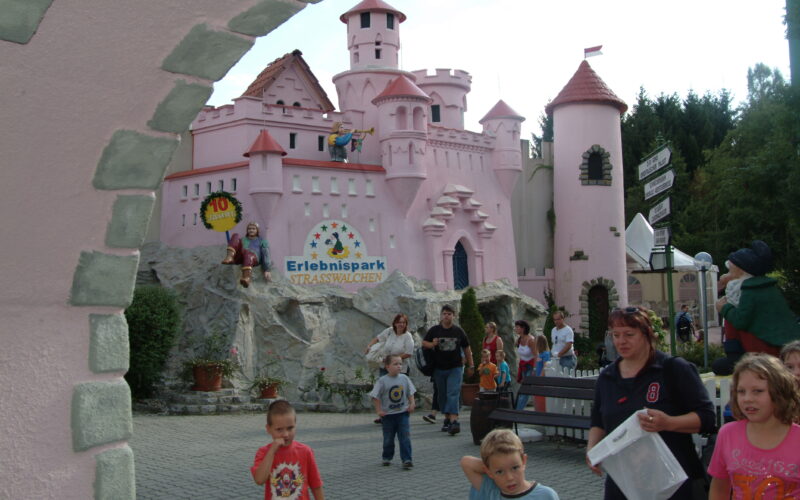 Menschen, darunter Kinder und Erwachsene, laufen und spielen in der Nähe eines rosafarbenen Märchenschlosses im Erlebnispark Strasswalchen. Im Hintergrund sind Schilder und Grünflächen zu sehen.