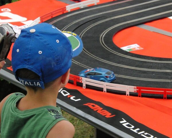 Ein Kind mit einer blauen Mütze beobachtet ein blaues Slotcar, das auf einer schwarzen Bahn bei einer Freiluftveranstaltung fährt.