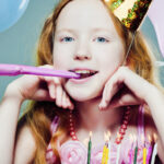 Ein Mädchen mit langen roten Haaren und einem Partyhut bläst in ein Partyhorn und sitzt vor einer Geburtstagstorte mit brennenden Kerzen und bunten Luftballons im Hintergrund.