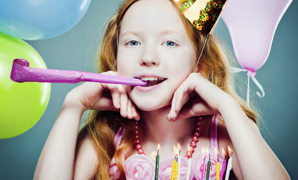 Ein Mädchen mit langen roten Haaren und einem Partyhut bläst in ein Partyhorn und sitzt vor einer Geburtstagstorte mit brennenden Kerzen und bunten Luftballons im Hintergrund.