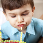Ein kleiner Junge in einem blauen Hemd bläst zwei brennende Kerzen auf einer Geburtstagstorte aus.