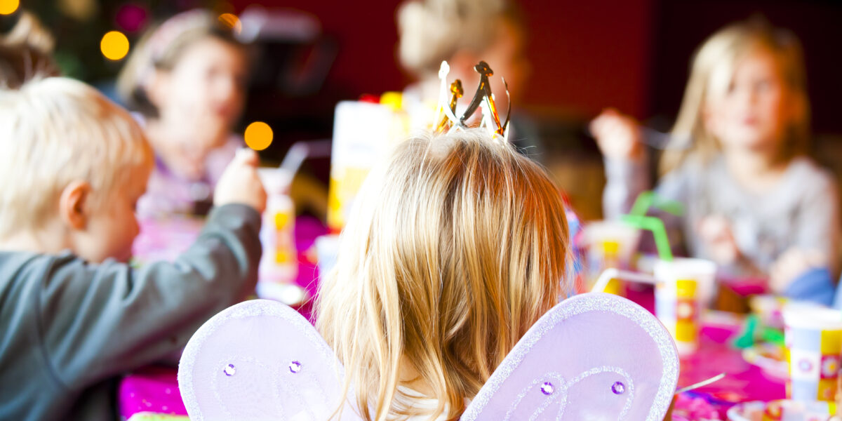 Eine Gruppe von Kindern sitzt auf einer Party um einen Tisch herum. Der Fokus liegt auf einem Kind, das Schmetterlingsflügel und eine Krone trägt, während andere im Hintergrund verschwommen sind.