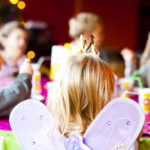 Eine Gruppe von Kindern sitzt auf einer Party um einen Tisch herum. Der Fokus liegt auf einem Kind, das Schmetterlingsflügel und eine Krone trägt, während andere im Hintergrund verschwommen sind.