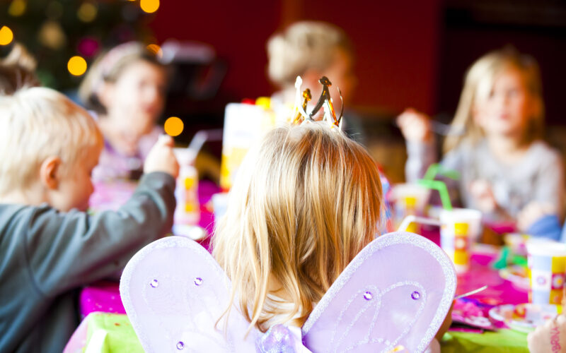 Eine Gruppe von Kindern sitzt auf einer Party um einen Tisch herum. Der Fokus liegt auf einem Kind, das Schmetterlingsflügel und eine Krone trägt, während andere im Hintergrund verschwommen sind.